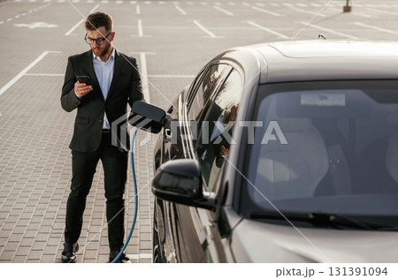 Holding phone in hand while vehicle is charging. Businessman in suit is near his black car outdoors Holding phone in hand while vehicle is charging. Businessman in suit is near his black car outdoors 131391094