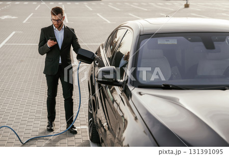 Holding phone in hand while vehicle is charging. Businessman in suit is near his black car outdoors 131391095