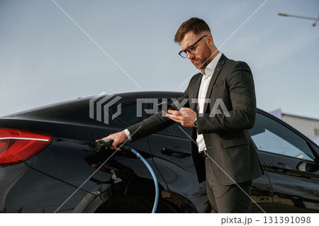 Holding phone in hand while vehicle is charging. Businessman in suit is near his black car outdoors Holding phone in hand while vehicle is charging. Businessman in suit is near his black car outdoors 131391098