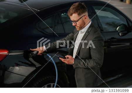 Green energy concept, charging. Businessman in suit is near his black car outdoors 131391099