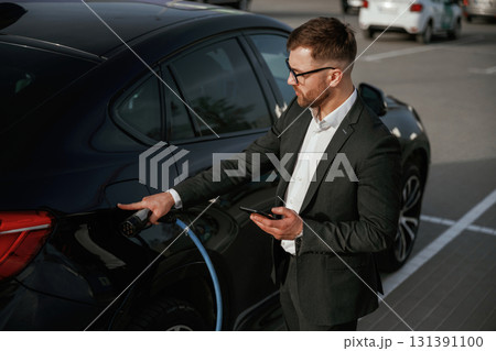 Green energy concept, charging. Businessman in suit is near his black car outdoors 131391100