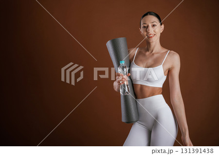 Mat and bottle of water in hands. Beautiful fitness woman is in the studio 131391438