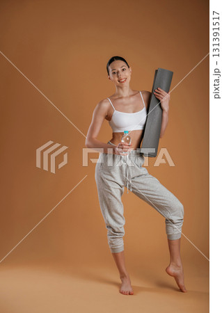 Looking forward, holding bottle of water and mat. Beautiful fitness woman is in the studio Looking forward, holding bottle of water and mat. Beautiful fitness woman is in the studio 131391517