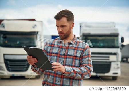 Having hard time reading the document in notepad. Man is standing against trucks outdoors Having hard time reading the document in notepad. Man is standing against trucks outdoors 131392360