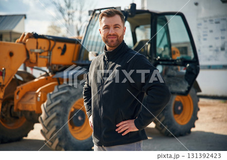 In black hoodie. Standing and posing. Man is with tractor. Agricultural worker In black hoodie. Standing and posing. Man is with tractor. Agricultural worker 131392423