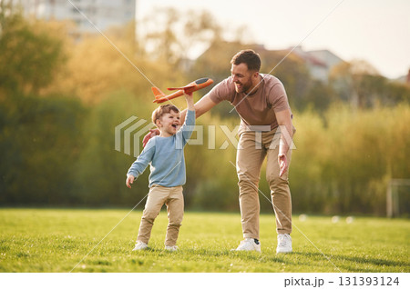 In casual clothes. Running with toy plane. Happy father with son are having fun on the field at summertime In casual clothes. Running with toy plane. Happy father with son are having fun on the field at summertime 131393124