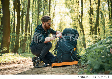 Looking for stuff in the backpack, sitting. Bearded man is in the forest at daytime 131393691