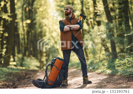 Yellow axe in hand, backpack on the ground. Bearded man is in the forest at daytime Yellow axe in hand, backpack on the ground. Bearded man is in the forest at daytime 131393761