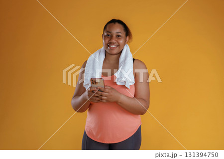 Holding smartphone, towel on the neck. African American woman is in the studio against yellow background Holding smartphone, towel on the neck. African American woman is in the studio against yellow background 131394750