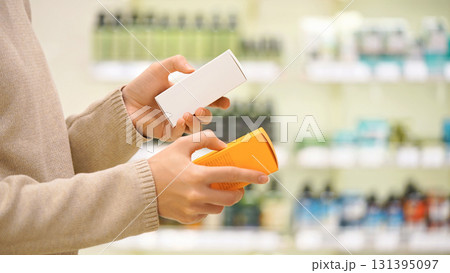 Close up of woman holding two cosmetic serums for face or hair in a beauty store, comparing products and reading labels. Female shopper choosing skincare or haircare item. Concept of conscious choice. 131395097