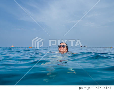 A young woman with sun glasses swimming in the blue sea. A blue hazy sky in the background A young woman with sun glasses swimming in the blue sea. A blue hazy sky in the background 131395121