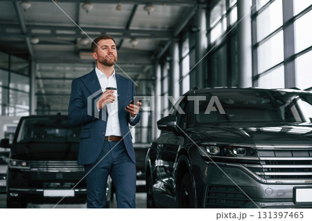 Break, with coffee and smartphone. A businessman is in a car dealership Break, with coffee and smartphone. A businessman is in a car dealership 131397465
