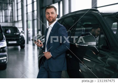 Leaning on the vehicle and holding smartphone. A businessman is in a car dealership 131397475