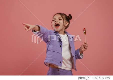 Showing to the side by finger. With candy. Cute little girl is against pink background 131397769