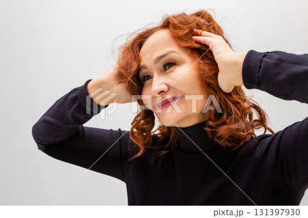beautiful 60 years old mature woman with curly red hair smiling confidently while touching her hair. 131397930