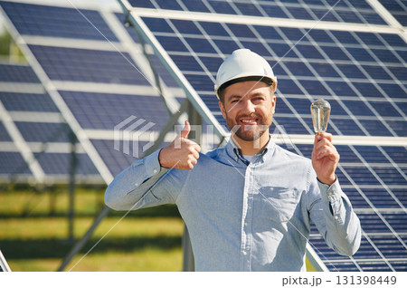 Smiling and holding bulb. Engineer with photovoltaic solar panels outdoors at daytime Smiling and holding bulb. Engineer with photovoltaic solar panels outdoors at daytime 131398449
