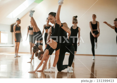Trainer is helping girl. Group of female kids practicing athletic exercises together indoors Trainer is helping girl. Group of female kids practicing athletic exercises together indoors 131399413