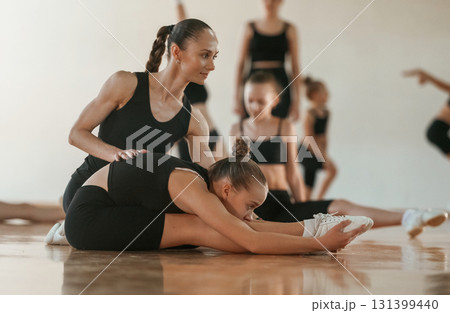 Woman with pigtail is helping little girl. Group of female kids practicing athletic exercises together indoors 131399440
