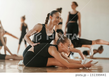 Woman with pigtail is helping little girl. Group of female kids practicing athletic exercises together indoors Woman with pigtail is helping little girl. Group of female kids practicing athletic exercises together indoors 131399441