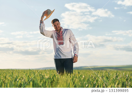 Taking off straw hat. Man in Ukrainian embroidery shirt is on the agricultural field at summer daytime Taking off straw hat. Man in Ukrainian embroidery shirt is on the agricultural field at summer daytime 131399895