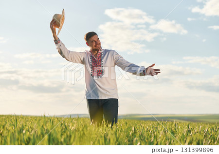 Taking off straw hat. Man in Ukrainian embroidery shirt is on the agricultural field at summer daytime Taking off straw hat. Man in Ukrainian embroidery shirt is on the agricultural field at summer daytime 131399896