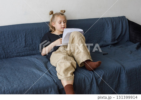 Happy little girl sitting on sofa reading book at home. Childhood and people concept 131399914