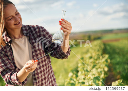 With test tube, doing research. Young woman is on the beautiful agricultural field at daytime With test tube, doing research. Young woman is on the beautiful agricultural field at daytime 131399987