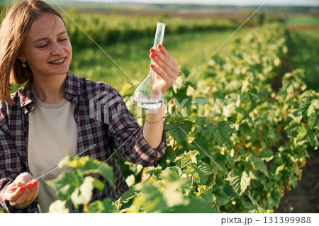 With test tube, doing research. Young woman is on the beautiful agricultural field at daytime With test tube, doing research. Young woman is on the beautiful agricultural field at daytime 131399988