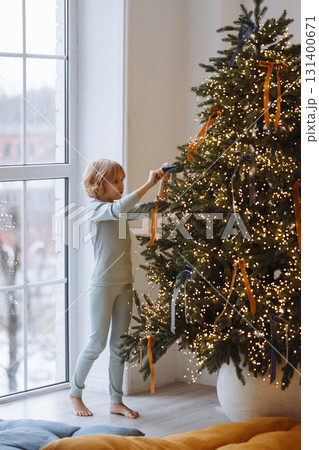Child decorating Christmas tree with ribbons in cozy room with white wall and large window Child decorating Christmas tree with ribbons in cozy room with white wall and large window 131400671