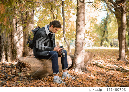 Young nature photographer sitting on log in forest, looking pictures after successful animals and nature photoshoot, satisfied with results 131400798