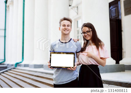 Cheerful students showing laptop with copy space and pointing to the screen, standing in front of university Cheerful students showing laptop with copy space and pointing to the screen, standing in front of university 131400811