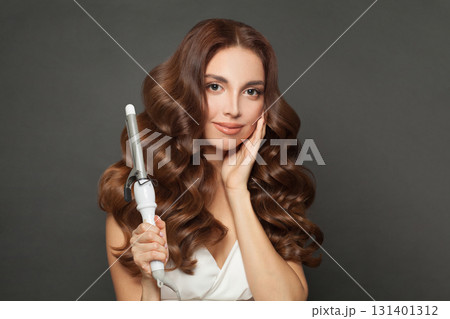 Fashionable woman with long wavy hairstyle using hair curling tongs iron, indoor portrait. 131401312