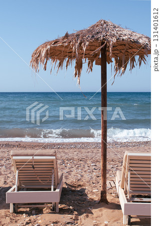 Simple beach scene on Crete - sunbeds and palm leaf umbrellas by the pebbles and sea 131401612