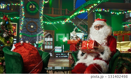 Amazed Santa Claus surprised to find gift box for him in cozy Christmas decorated room. Elderly man playing Santa Claus astonished after finding present on table, Christmas miracle concept, camera A 131402498