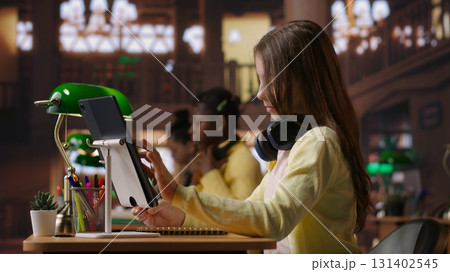 Preteen student using a tablet to complete her class notes in the library, uses he digital academic database to consult learning materials. Preparing diligently to pass her exams. Camera B. 131402545