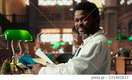 African american medical student reads under green lamps in a library, doing research about anatomy materials and case files. Exam preparation highlights the commitment in medical school. Camera B. African american medical student reads under green lamps in a library, doing research about anatomy materials and case files. Exam preparation highlights the commitment in medical school. Camera B. 131402617