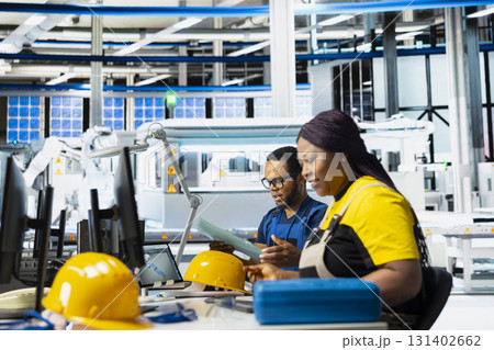 Black engineer in solar panel factory reviewing photovoltaic system performance on laptop. Female employee analyzing production line to optimize efficiency in industrial manufacturing plant. 131402662