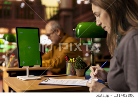 Mature woman collecting notes for her thesis next to green screen at the campus library, writing down ideas and finding reliable citation sources for her paperwork. Academic pursuit. 131402806