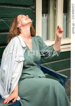 Woman in a green dress with a denim jacket over her shoulder leans back on a wooden bench, smiling and enjoying the sunlight 131406457