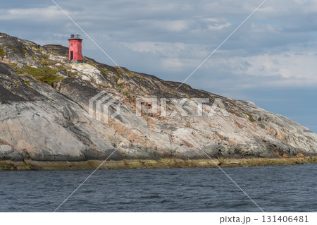 A small red lighthouse stands on rugged rocky cliffs above dark blue water under cloudy skies A small red lighthouse stands on rugged rocky cliffs above dark blue water under cloudy skies 131406481