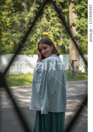 Portrait of a woman in a light denim jacket standing outdoors near a white wooden fence, looking over her shoulder thoughtfully Portrait of a woman in a light denim jacket standing outdoors near a white wooden fence, looking over her shoulder thoughtfully 131406483