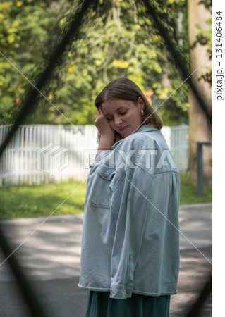 Portrait of a woman in a light denim jacket standing outdoors near a white wooden fence, looking over her shoulder thoughtfully Portrait of a woman in a light denim jacket standing outdoors near a white wooden fence, looking over her shoulder thoughtfully 131406484