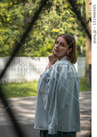 Portrait of a woman in a light denim jacket standing outdoors near a white wooden fence, looking over her shoulder thoughtfully Portrait of a woman in a light denim jacket standing outdoors near a white wooden fence, looking over her shoulder thoughtfully 131406485