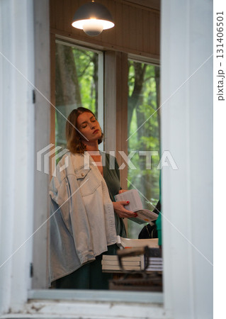 Woman in a green dress and denim jacket stands by a window, holding a book with stacked volumes behind her 131406501