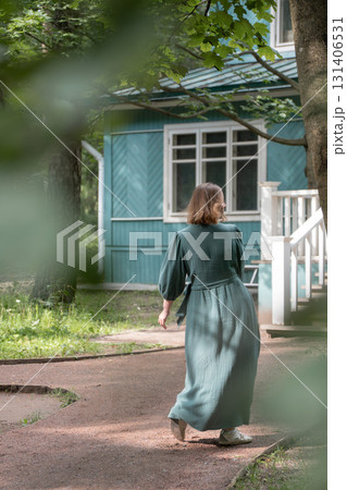 A woman in a long green dress walks along a garden path near a wooden house, partly framed by leaves 131406531