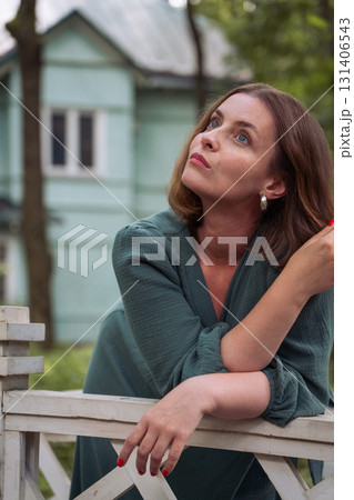 A woman in a dress leans thoughtfully on a white wooden fence, gazing upward with an expressive look against the backdrop of a turquoise country house 131406543