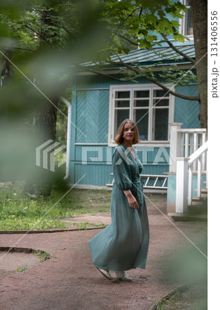 A woman in a flowing green dress spins gracefully on a garden path in front of a wooden house 131406556