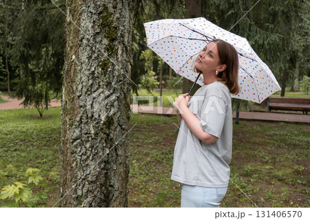 Woman with short hair in a gray t-shirt holds a white umbrella with colorful dots in a green park in Peredelkino 131406570