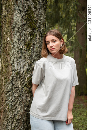 Woman in a light gray t-shirt leans against a tree, looking down thoughtfully with a calm expression 131406594