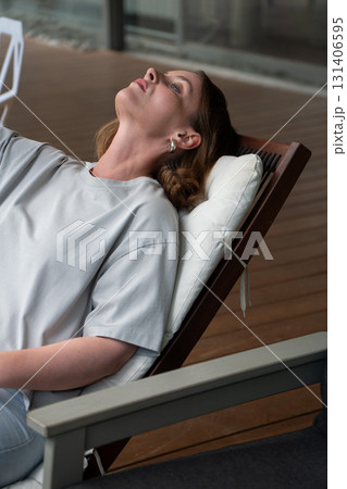 A woman in a light gray t-shirt reclines on a cushioned wooden lounge chair, gazing upward in a calm, reflective mood A woman in a light gray t-shirt reclines on a cushioned wooden lounge chair, gazing upward in a calm, reflective mood 131406595